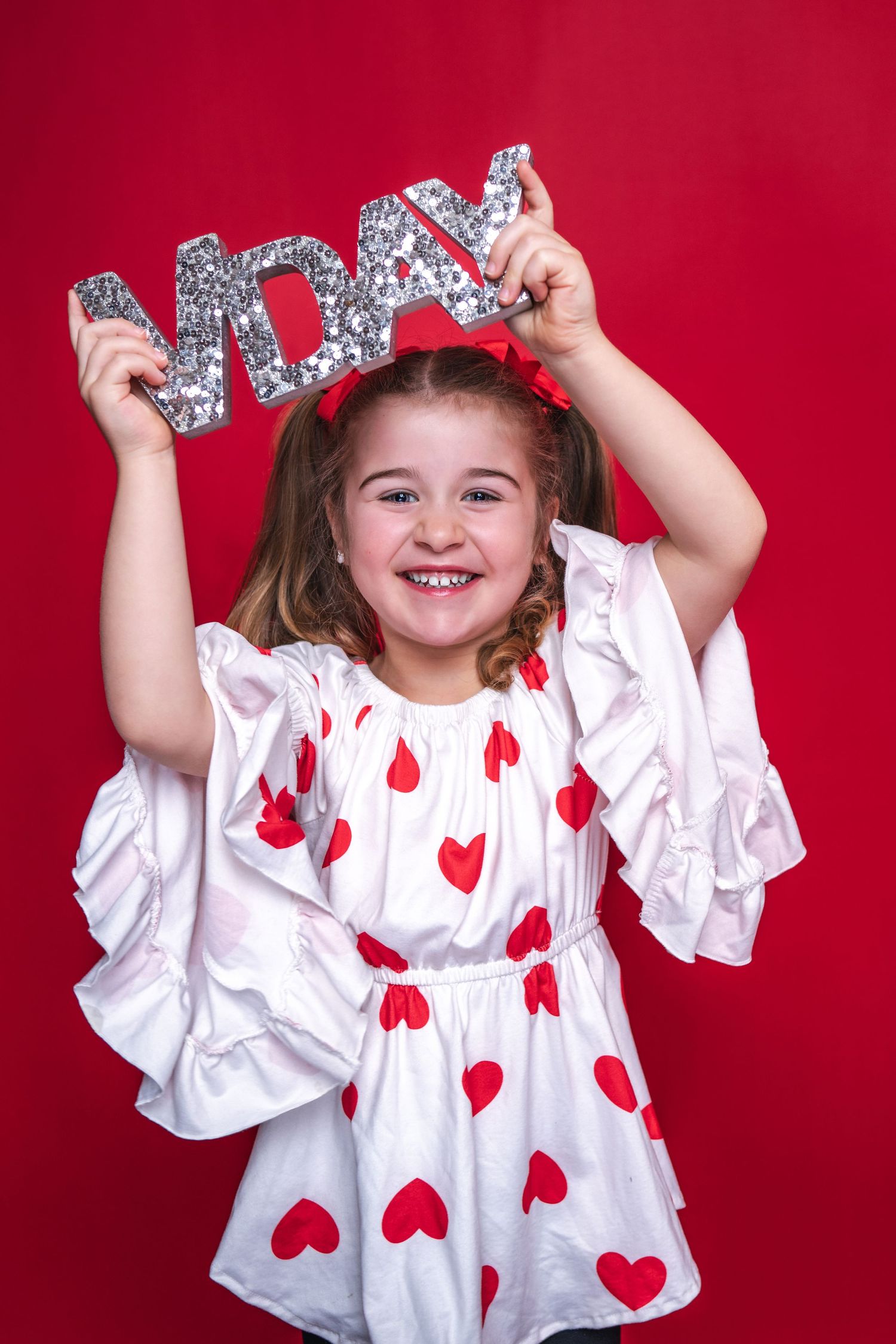 Sparkling silver LOVE text held up against red backdrop with white dress decorated in red heart pattern.