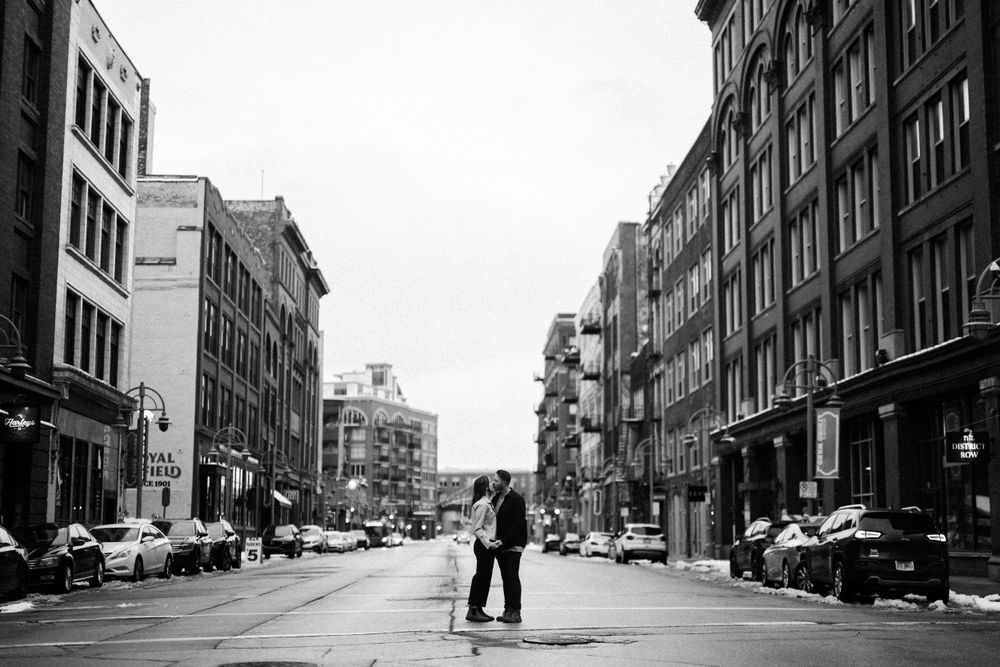 A black and white urban street scene with figures embracing in the center, surrounded by historic buildings and parked cars.
