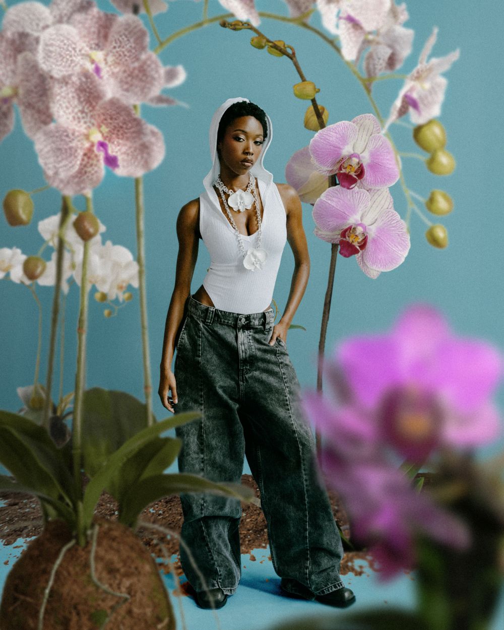 Fashion model posing against turquoise background with pink orchids and decorative elements.