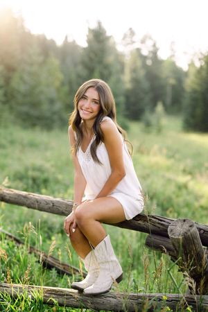 Smiling young woman in white dress and cowboy boots sitting on rustic wooden fence in sunny green meadow.