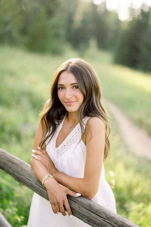 Young woman in white sundress leaning on wooden fence rail in lush green meadow, smiling softly at camera.