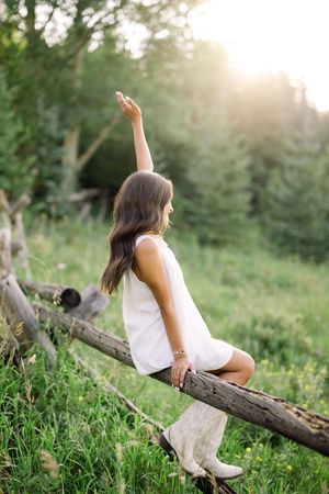 Young woman in white dress and boots sits on a rustic log fence, raising her arm in a sunlit green meadow.