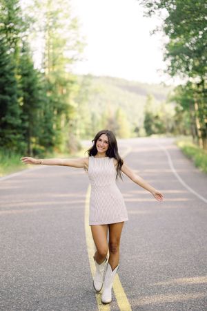 Young woman in white mini dress and cowboy boots posing on a tree-lined road with arms outstretched, smiling.