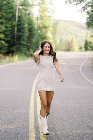 Young woman in a mini dress and white cowboy boots walks confidently down a tree-lined road, smiling.