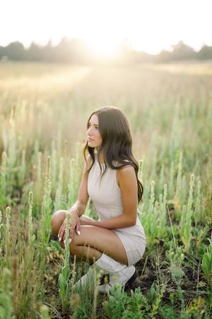 Young woman in beige dress crouching in a sunlit green field during golden hour.