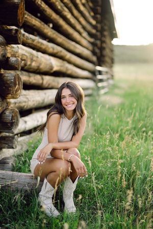 Young woman in white dress and cowboy boots crouching by rustic log cabin wall in green meadow, smiling.