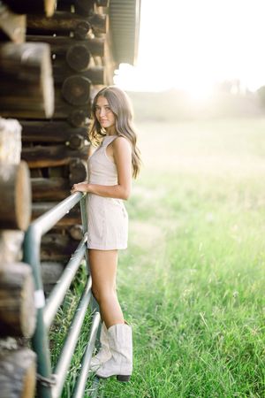 Young woman in white romper and cowboy boots poses by a rustic log cabin in a sunlit green field.