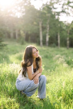 Young woman with long dark hair crouching in a sunlit green meadow, wearing jeans and a white top.