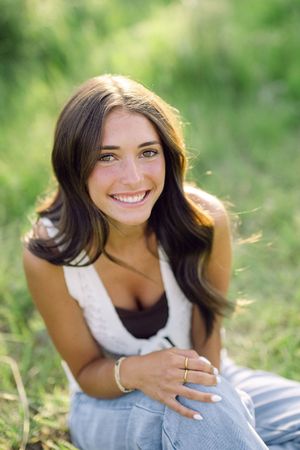 Smiling brunette woman sitting on green grass outdoors, wearing white top and jeans, with gold bracelet and ring.