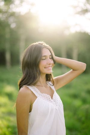 Young woman in white sundress smiling peacefully outdoors with eyes closed, bathed in golden sunlight among green trees.