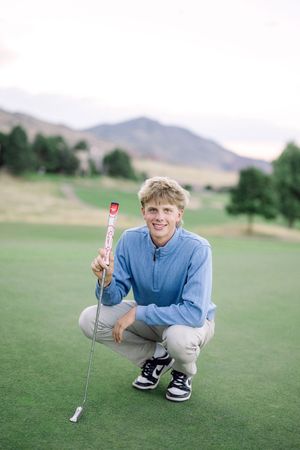 A golfer crouches on a green course examining the terrain with mountains visible in the background.