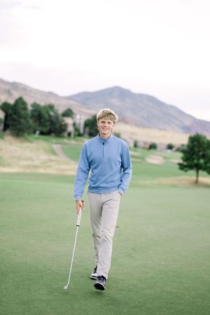 Golfer in blue sweater and khaki pants stands on putting green with mountains in background.