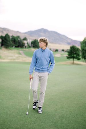 Golfer in blue quarter-zip sweater and khaki pants walking on green course with mountains in background.