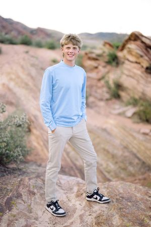 A person wearing a light blue sweater and khaki pants poses on rocky desert terrain at sunset.