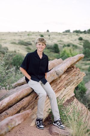 Person in black polo shirt and khaki pants sitting on red rocks at Red Rocks Park in Colorado during daytime.