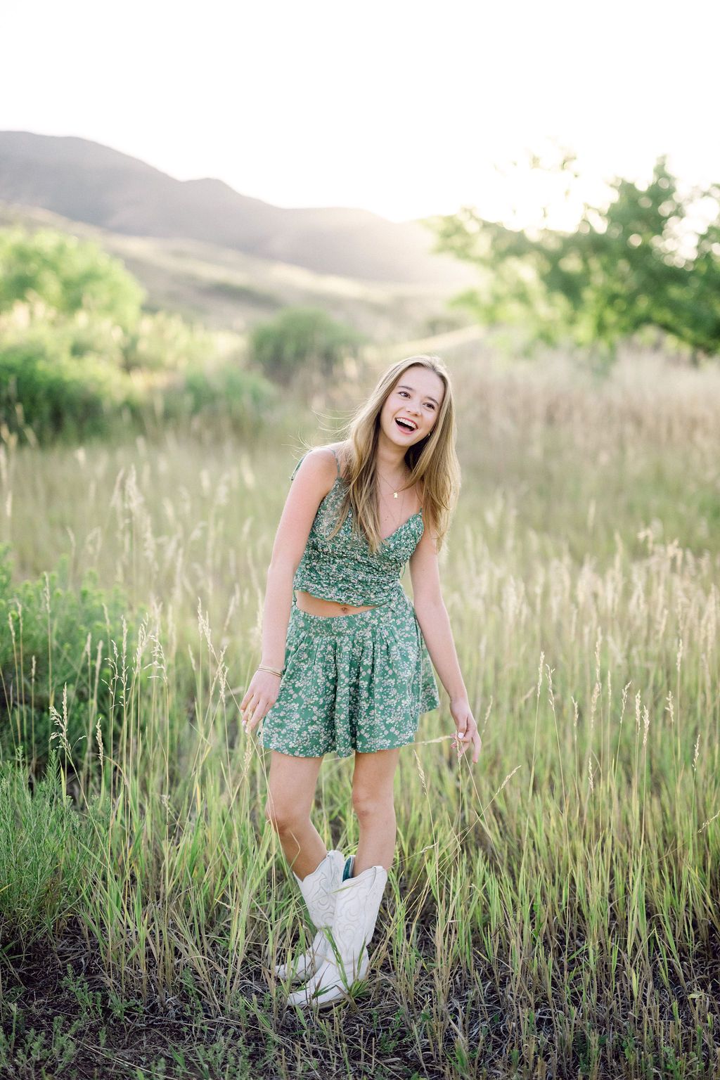 A person in a floral green dress and white shoes laughs joyfully while standing in a sunlit meadow of tall grass.