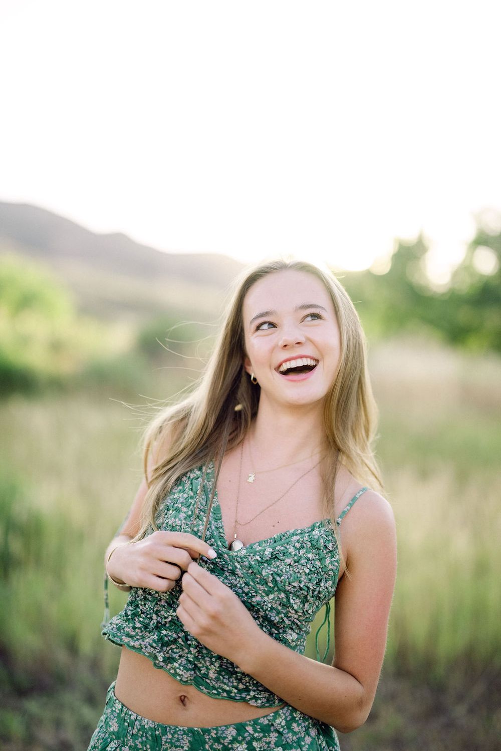 A person in a green floral dress poses outdoors in a grassy field on a sunny day.