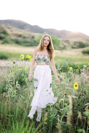 A person in a flowing white dress dances gracefully among wildflowers and tall grass in a mountain meadow.