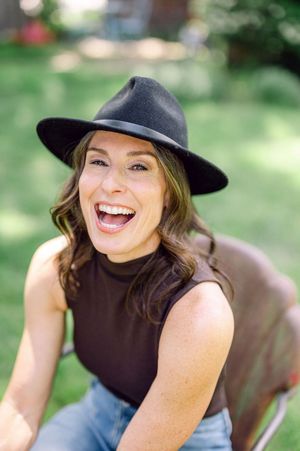 A laughing woman wearing a black wide-brim hat and dark sleeveless top sits outdoors in a sunny green garden.