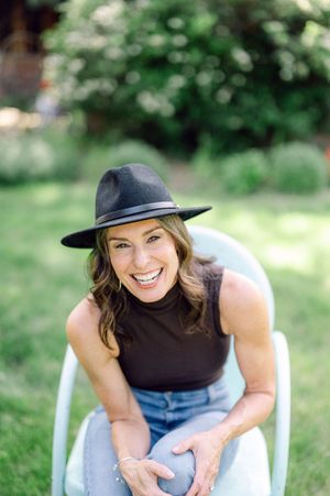 Smiling woman in black top, jeans, and wide-brim hat sits outdoors in white chair with lush green garden background.