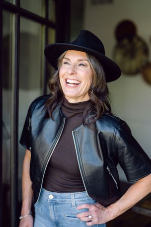 Smiling woman wearing a black wide-brim hat and leather vest over a turtleneck with jeans, posing confidently indoors.