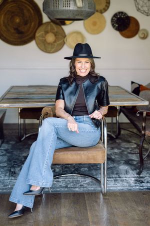 Stylish woman in black hat, leather vest, and wide-leg jeans seated in modern chair with woven wall decor.