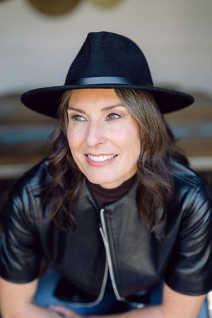 Smiling woman wearing a black wide-brim hat and leather jacket, posing casually in a bright outdoor setting.