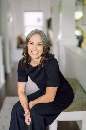 Smiling woman with gray-streaked hair wearing a black dress, sitting in a bright modern hallway interior.