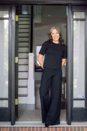 Woman in black outfit standing confidently in open glass doorway of elegant home, smiling warmly.