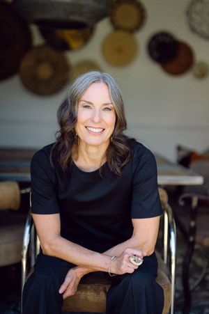 Smiling woman with gray-streaked hair in black dress seated in modern chair with decorative wall art behind her.