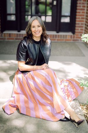 Woman smiling in black leather top and pink striped maxi skirt seated outdoors near brick building.