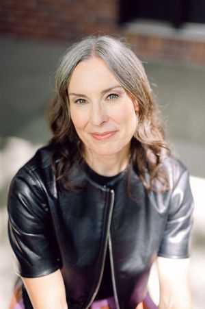 Woman in black leather top with gray-streaked hair smiling warmly outdoors in a candid portrait photo.