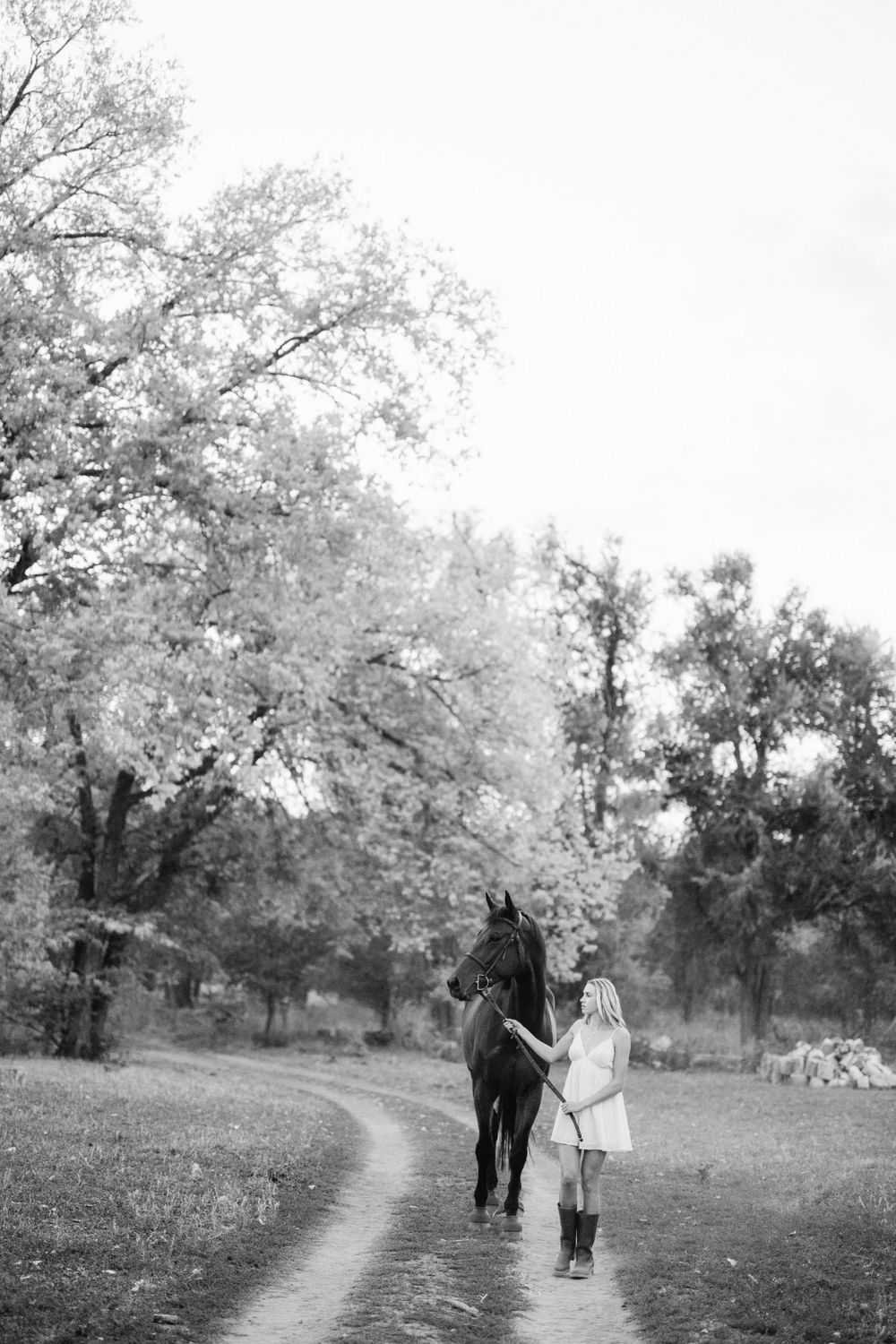 A black and white photo showing someone walking with their horse down a tree-lined path.