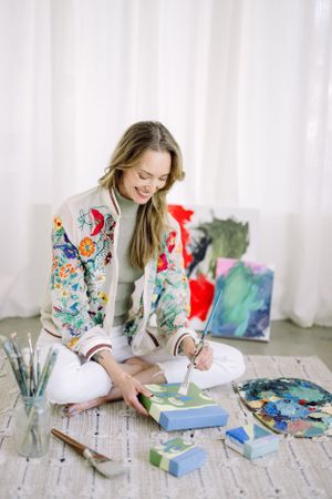 Artist in colorful floral jacket working on painting while sitting on floor with art supplies.