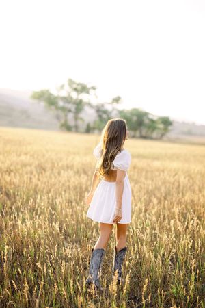 Young woman in white dress and cowboy boots standing in golden field at sunset.