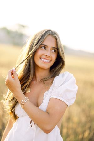 Smiling young woman in white dress holding hair in golden field at sunset.