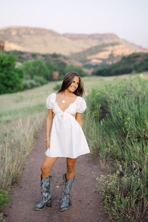 Young woman in white mini dress and cowboy boots standing on a dirt path in a scenic green meadow with mountains.
