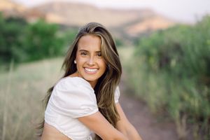Smiling young woman with long brown hair in a white top posing outdoors on a scenic trail.