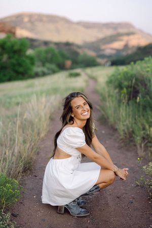 Young woman in white crop top and skirt crouching on a dirt path, smiling, with green fields and mountains behind her.