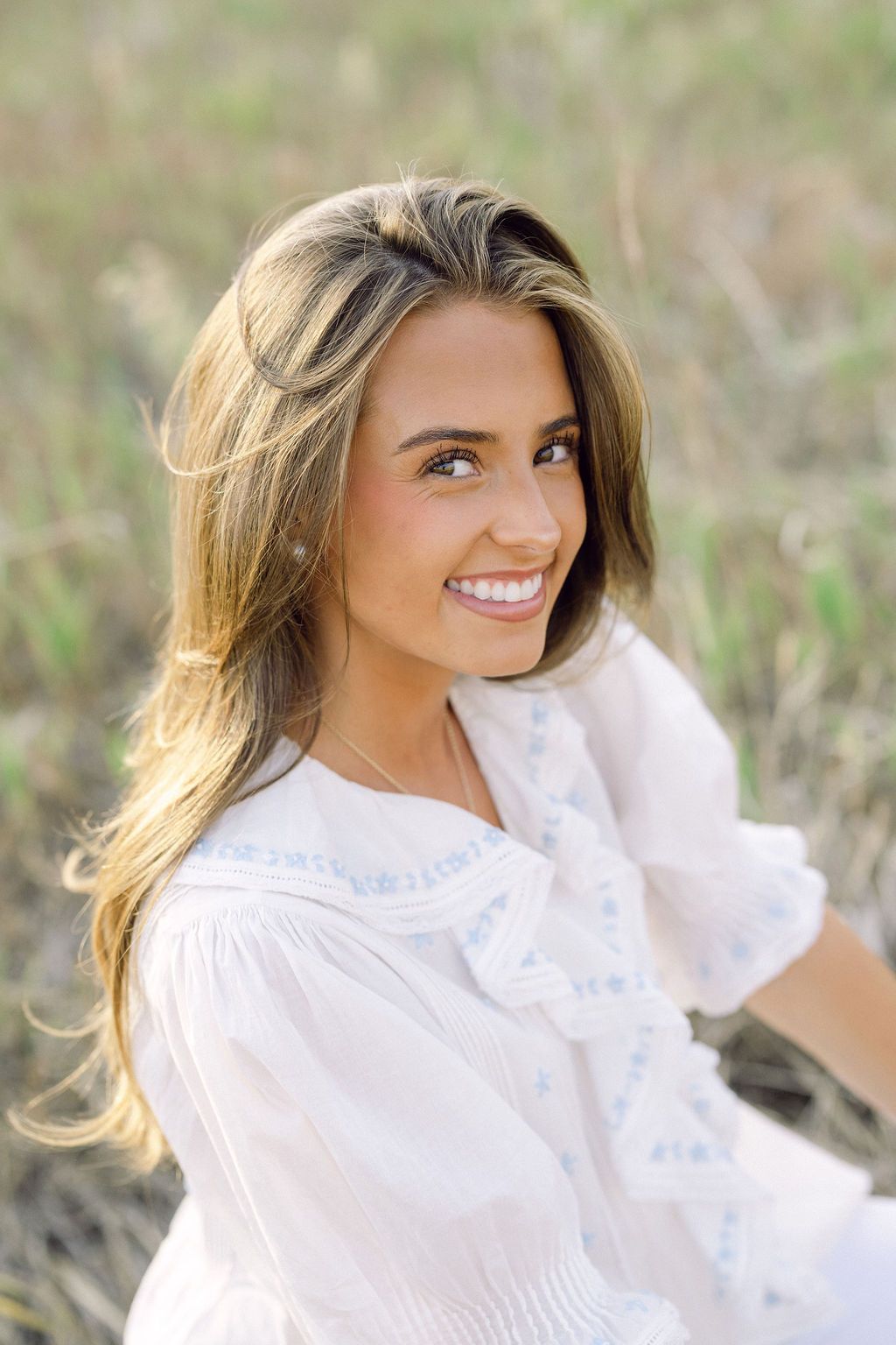 Young woman with long blonde hair smiling outdoors in a white embroidered blouse, sitting in a sunlit grassy field.