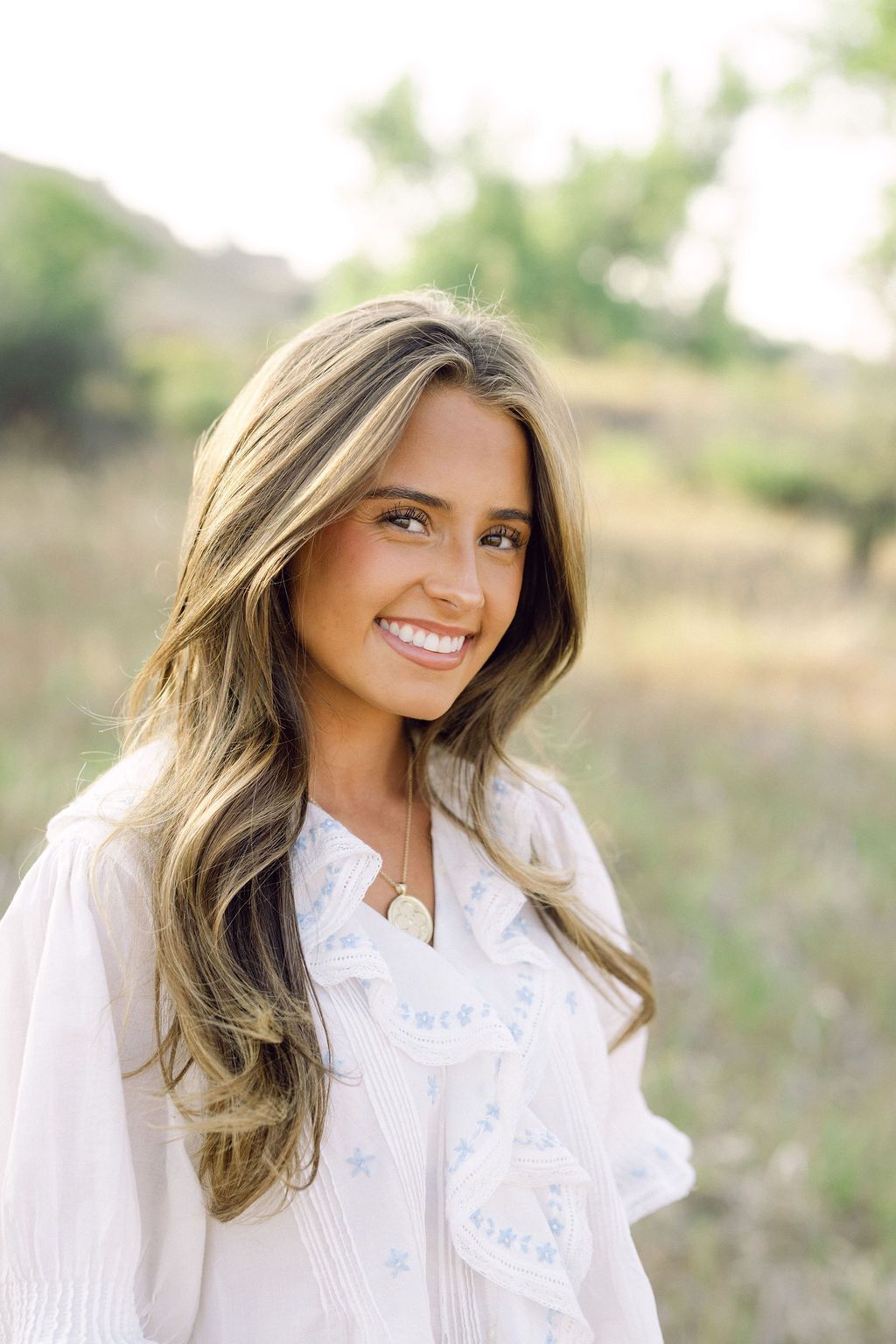 Smiling young woman with long highlighted hair wearing a white embroidered blouse outdoors in a sunny field.