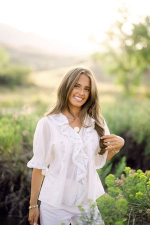 Smiling young woman in white ruffled blouse standing in a sunlit green field with mountains in the background.