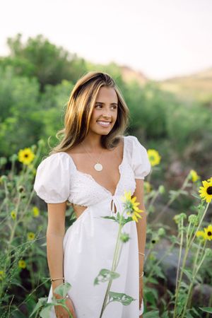 Smiling woman in white dress among sunflowers in a green field, wearing a gold necklace.