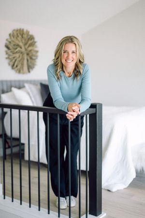Someone wearing a light blue sweater and dark pants leans casually against a black stair railing in a bright bedroom.