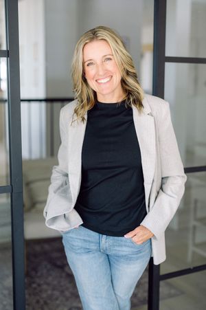 A woman wearing a black top and light gray blazer with blue jeans stands casually against a wall in an indoor setting.
