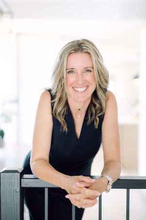 Professional portrait of someone in a sleeveless black top leaning on a railing with a warm smile in a bright interior setting.