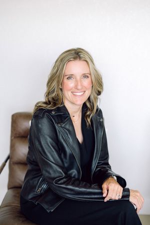 Professional portrait of someone wearing a black leather jacket and black top seated on a brown leather chair.