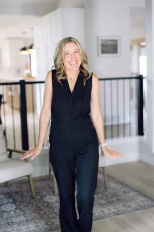 Someone wearing a sleek black sleeveless jumpsuit stands smiling in a modern interior space with white walls and gray rug.