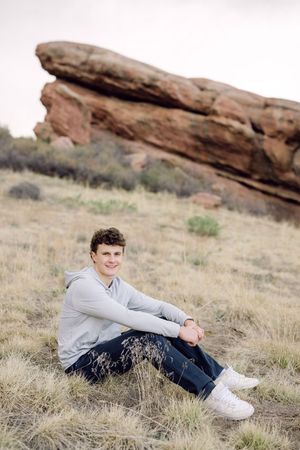 Young man sitting on grassy hillside at Red Rocks Colorado, wearing gray hoodie, jeans, and white sneakers, smiling at camera.