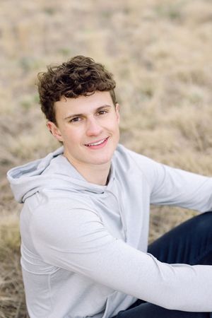 Young man with curly hair wearing a gray hoodie sitting outdoors in a field, smiling at the camera.